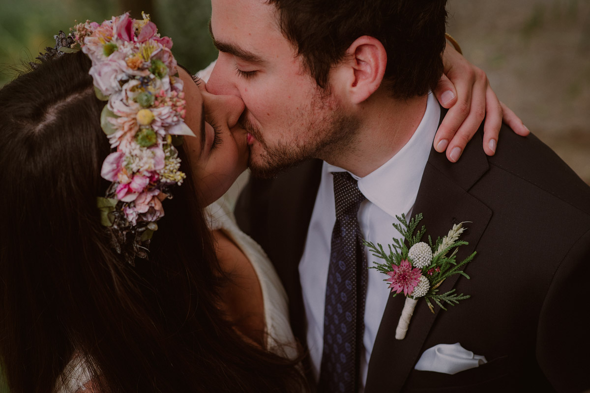Fotógrafo en Donostia - San Sebastian, Gipuzkoa | La pareja se besa en su reportaje fotográfico de bodas. Ella luce una corona de flores naturales en tonos rosados y verdes a juego con la flor de la solapa del novio.